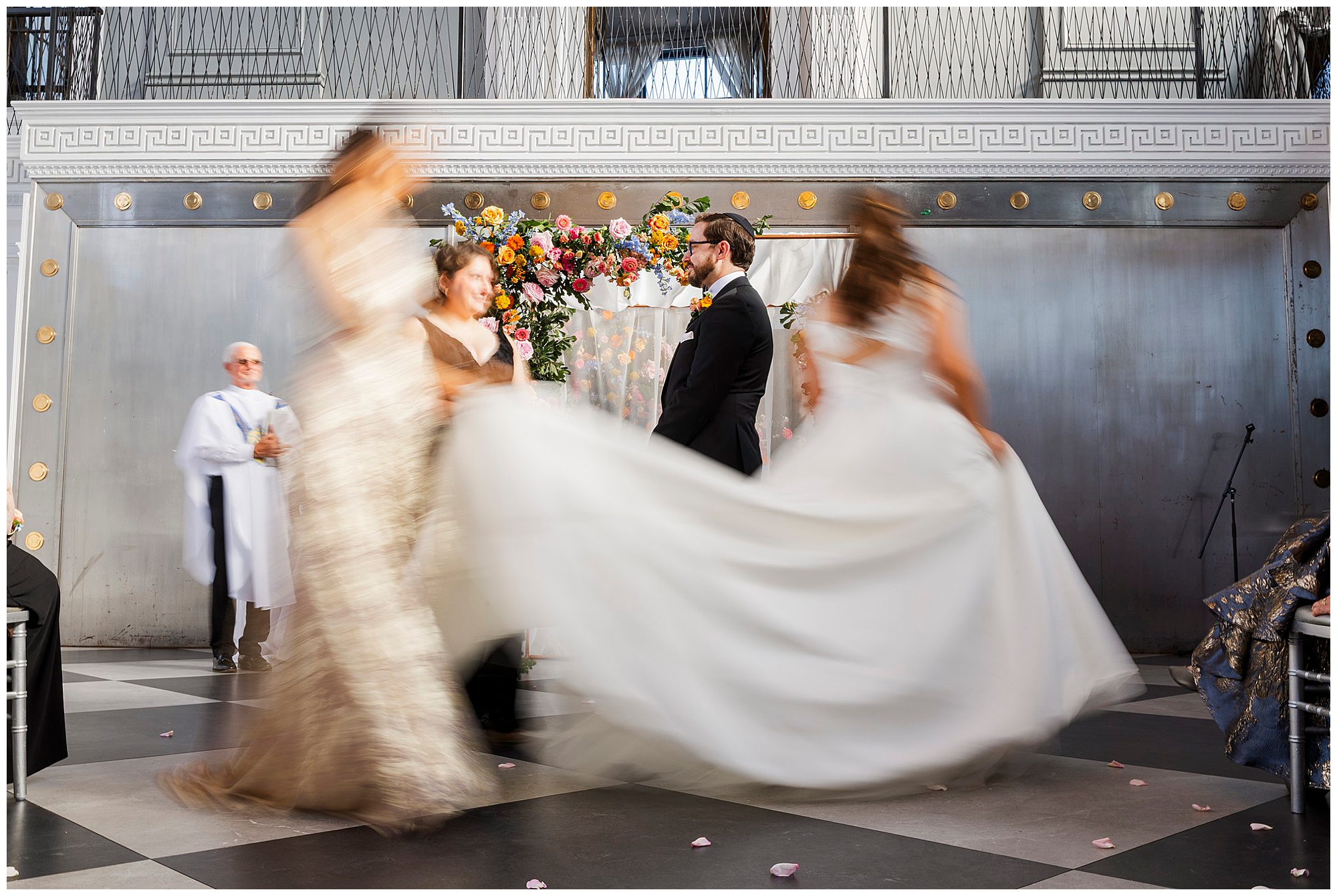 A felina wedding ceremony scene with a bride and groom under a floral arch. Two women in dresses move, creating a motion blur, while an officiant stands nearby. The background features metallic and classical decor.