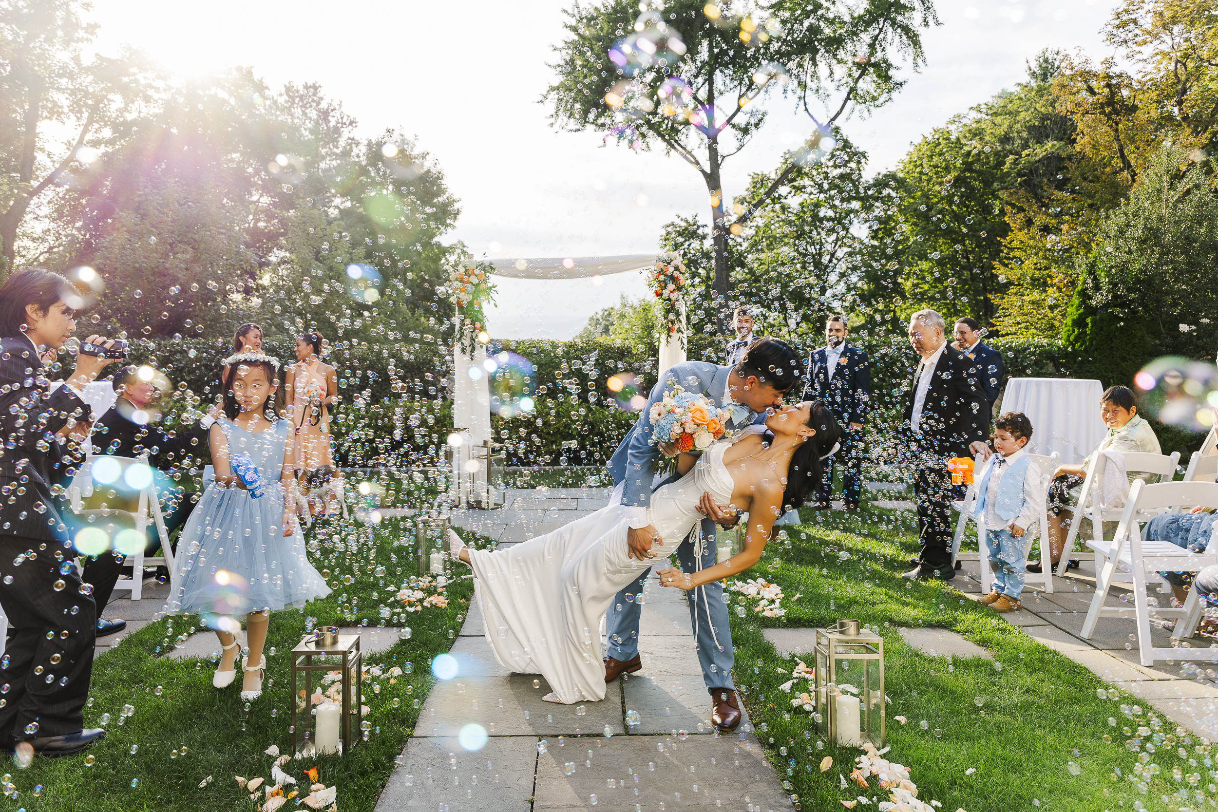 A bride and groom kiss as the groom dips the bride in an outdoor wedding ceremony, surrounded by guests blowing bubbles, with flower petals on the ground and sunlight streaming through the trees.