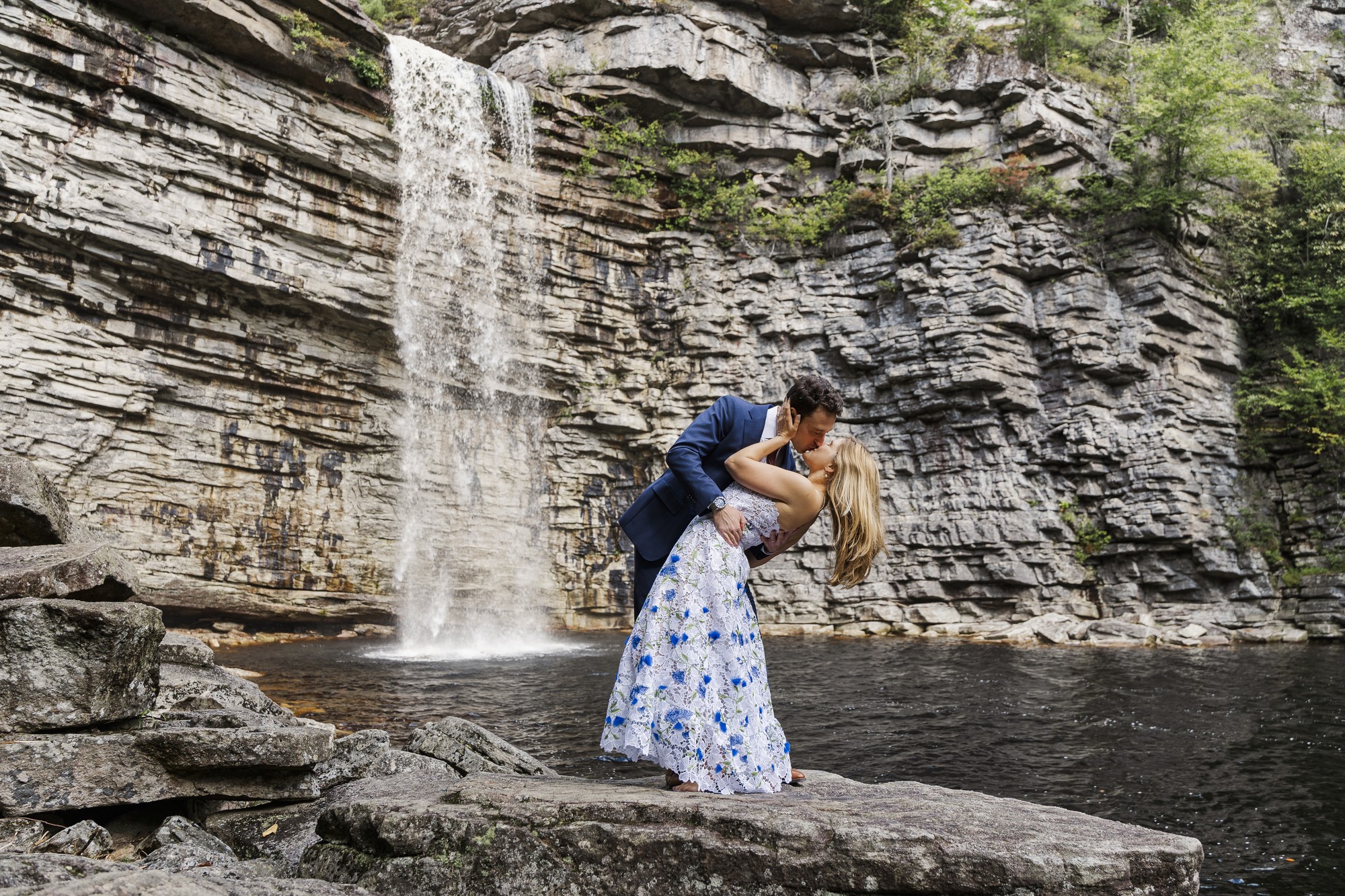 A man in a suit dips and kisses a woman in a white floral dress on a rock beside a waterfall at their Lake Minnewaska elopement, surrounded by rocky cliffs and lush greenery.