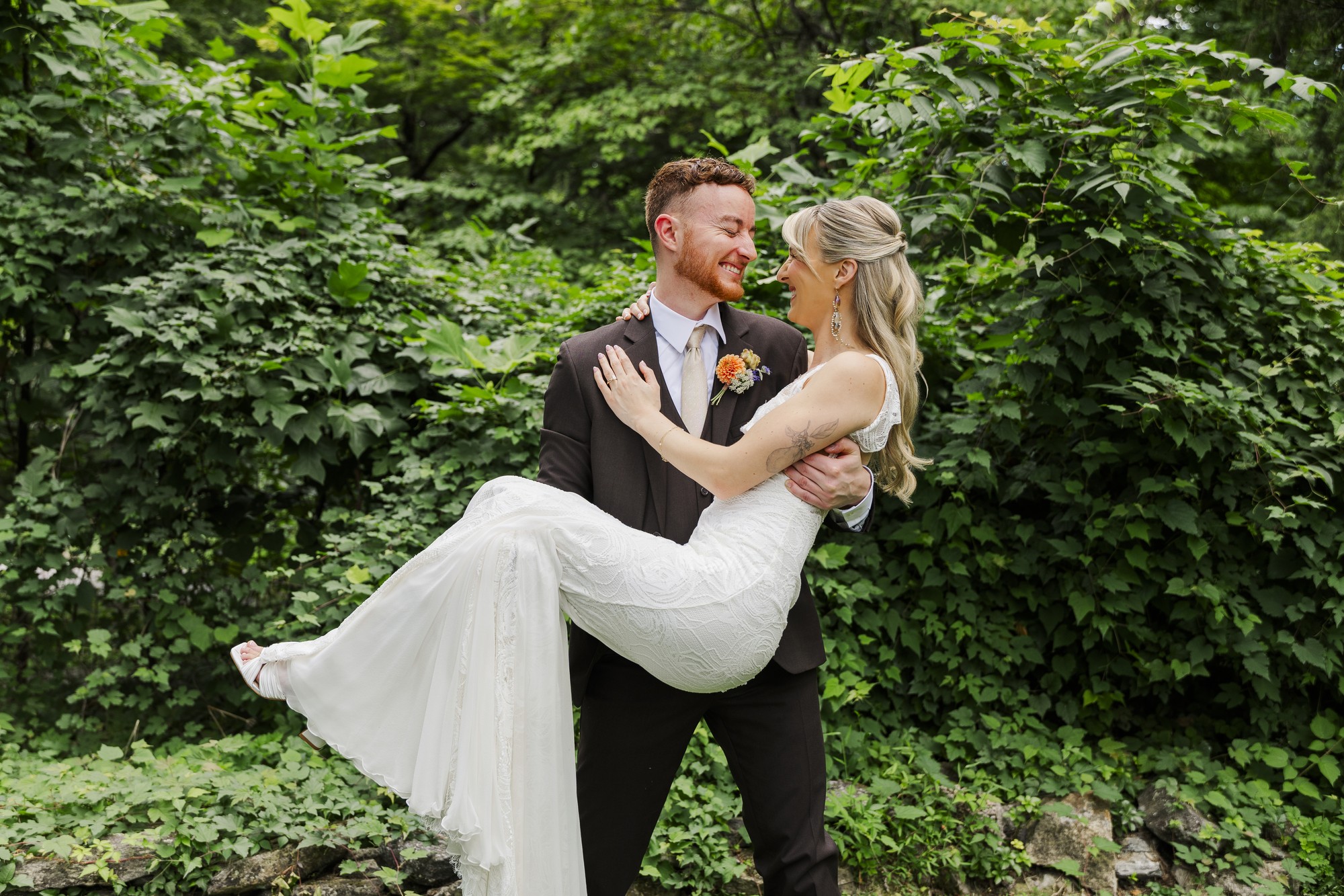A groom in a black suit lifts and smiles at his bride in a white dress outdoors, surrounded by lush green foliage during their picturesque Poughkeepsie area wedding at a historic estate.