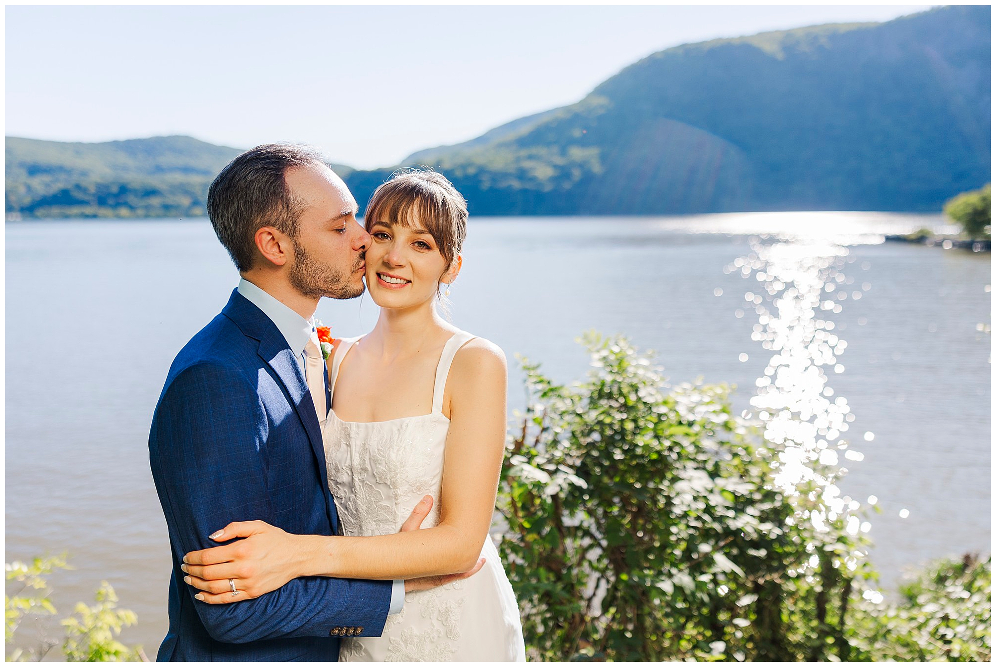 Groom in a blue suit kisses his bride's cheek as they embrace beside the sparkling river and green hills.