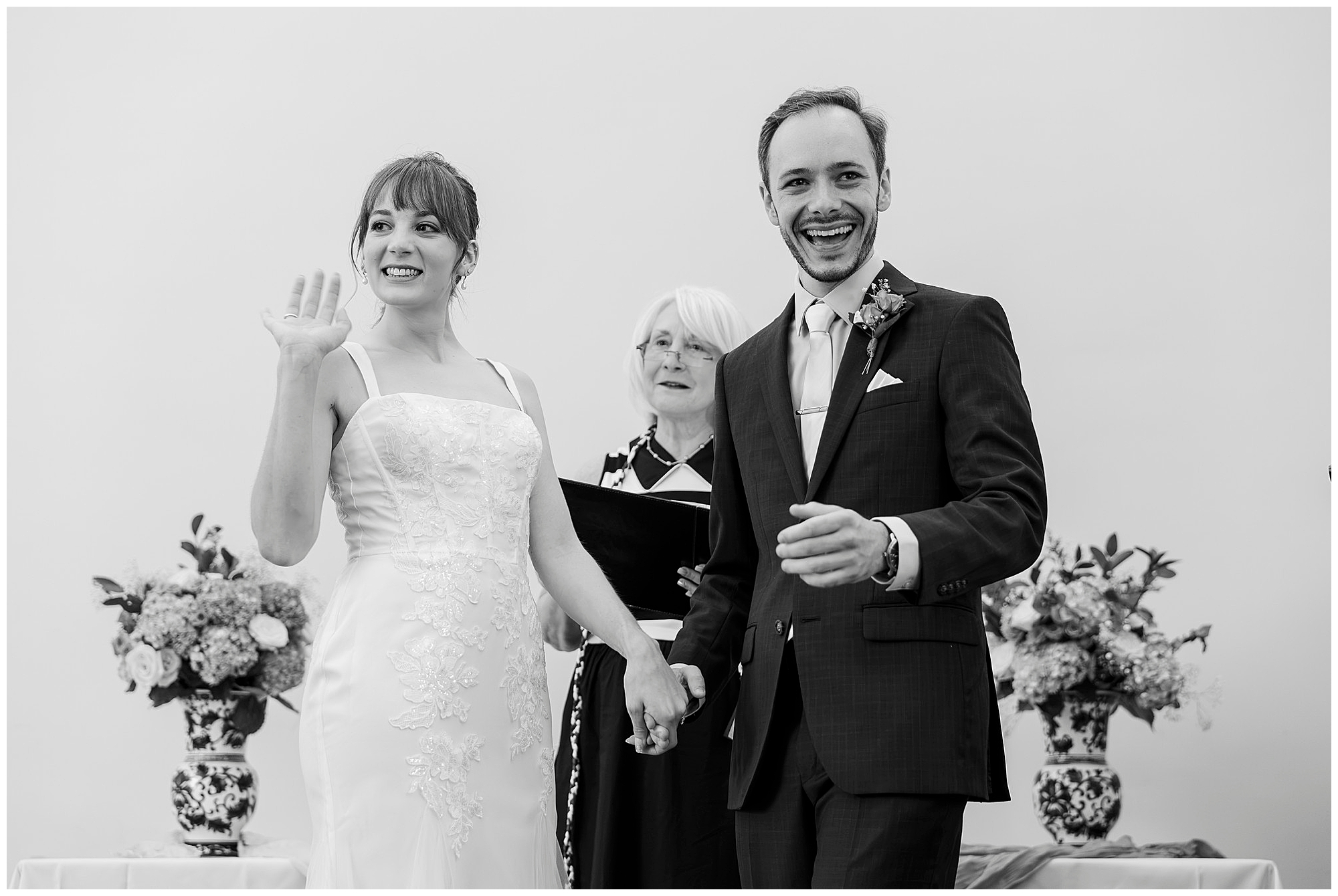 Bride waves and smiles while holding the groom's hand, the officiant behind them, in black and white.