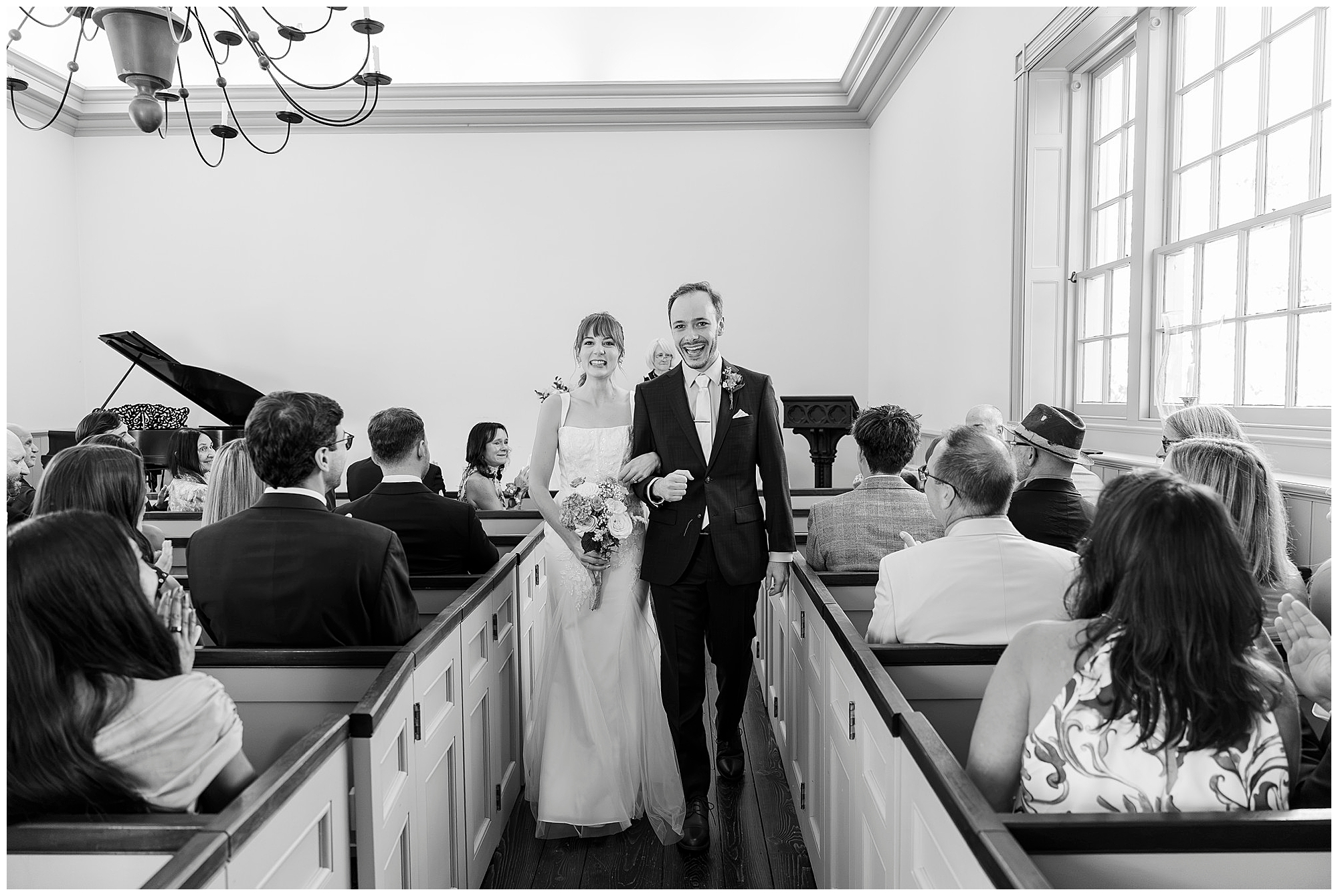 Smiling bride and groom walk back up the aisle arm in arm with guests seated on both sides of the small, bright chapel.