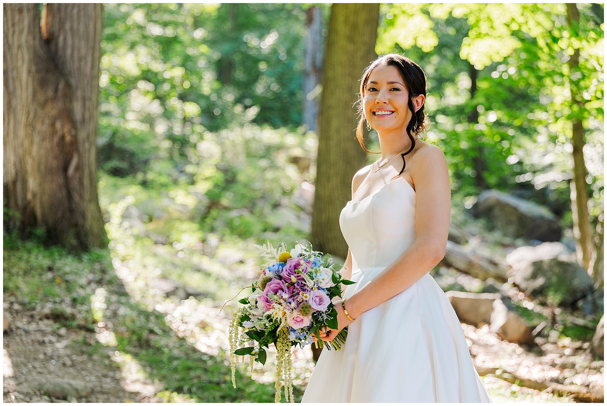 Bride in a white wedding dress smiles in a sunlit forest, holding a bouquet of purple and white flowers.