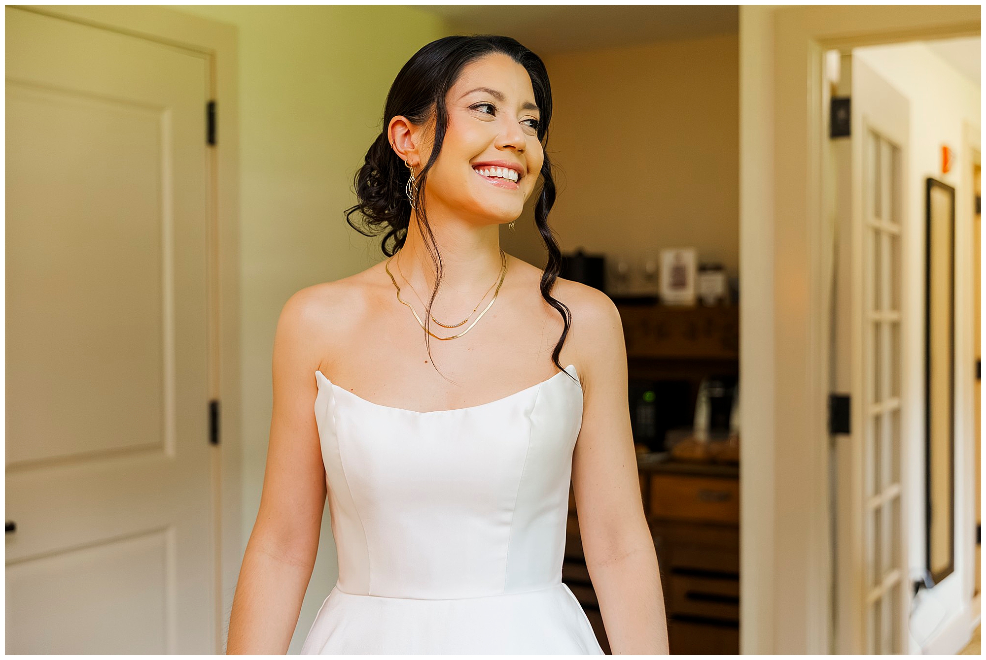 Smiling bride in a white strapless dress with a curled updo and gold jewelry looks to the side, doors and wooden furniture behind her.