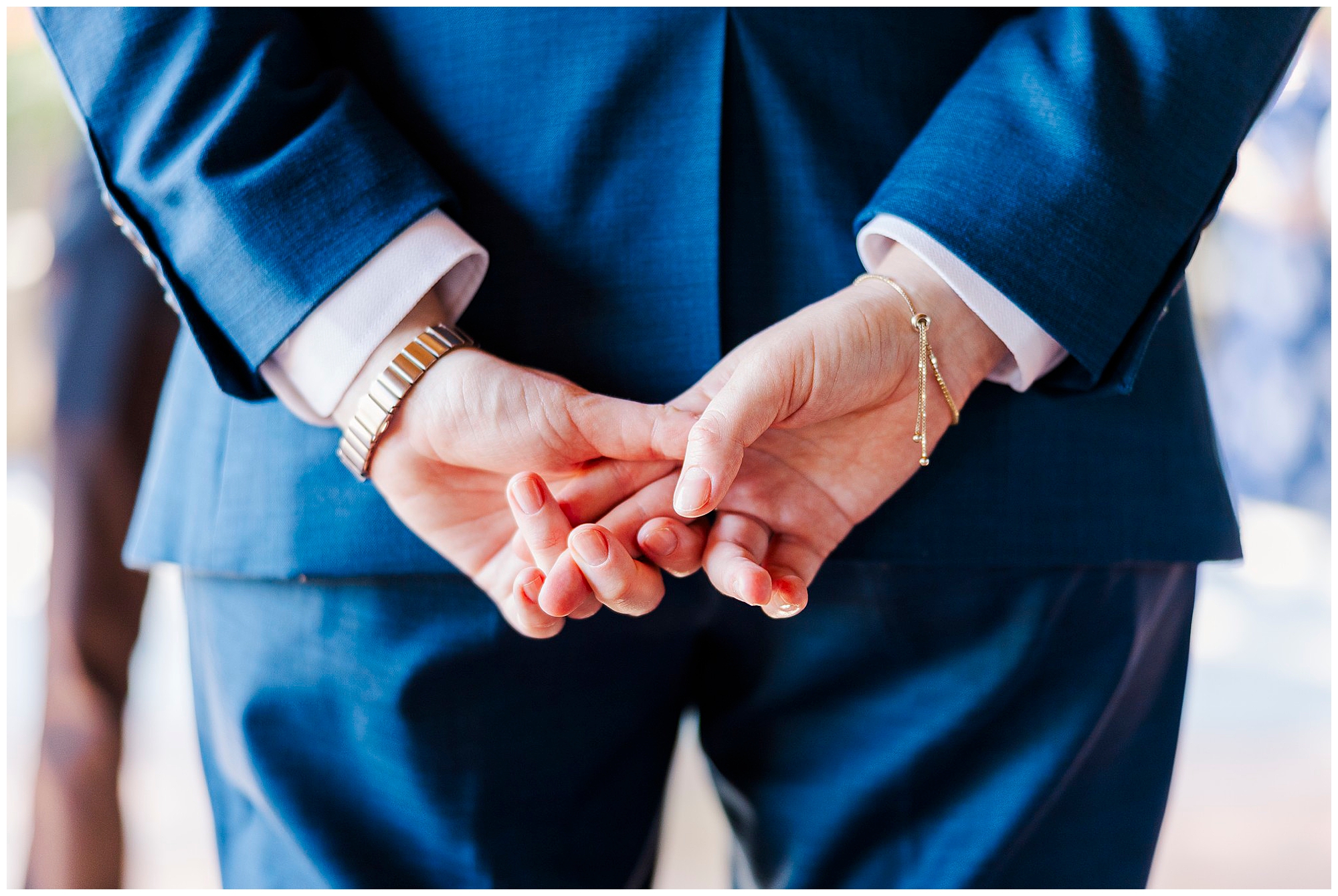 Close-up of a bride in a blue suit with her hands clasped behind her back, wearing a watch and a delicate bracelet.