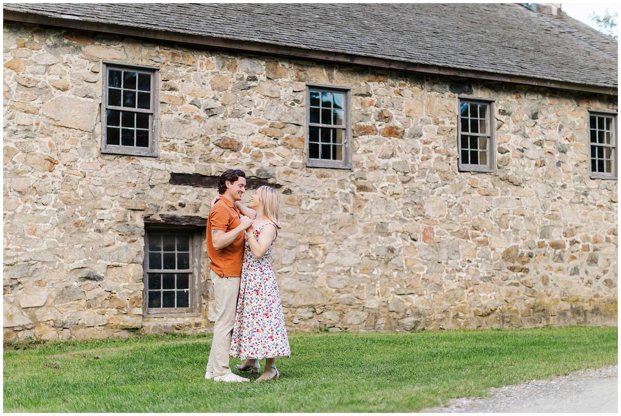 Couple smiles and holds hands on the lawn in front of a historic stone building during their engagement photos at Waterloo Village in New Jersey.