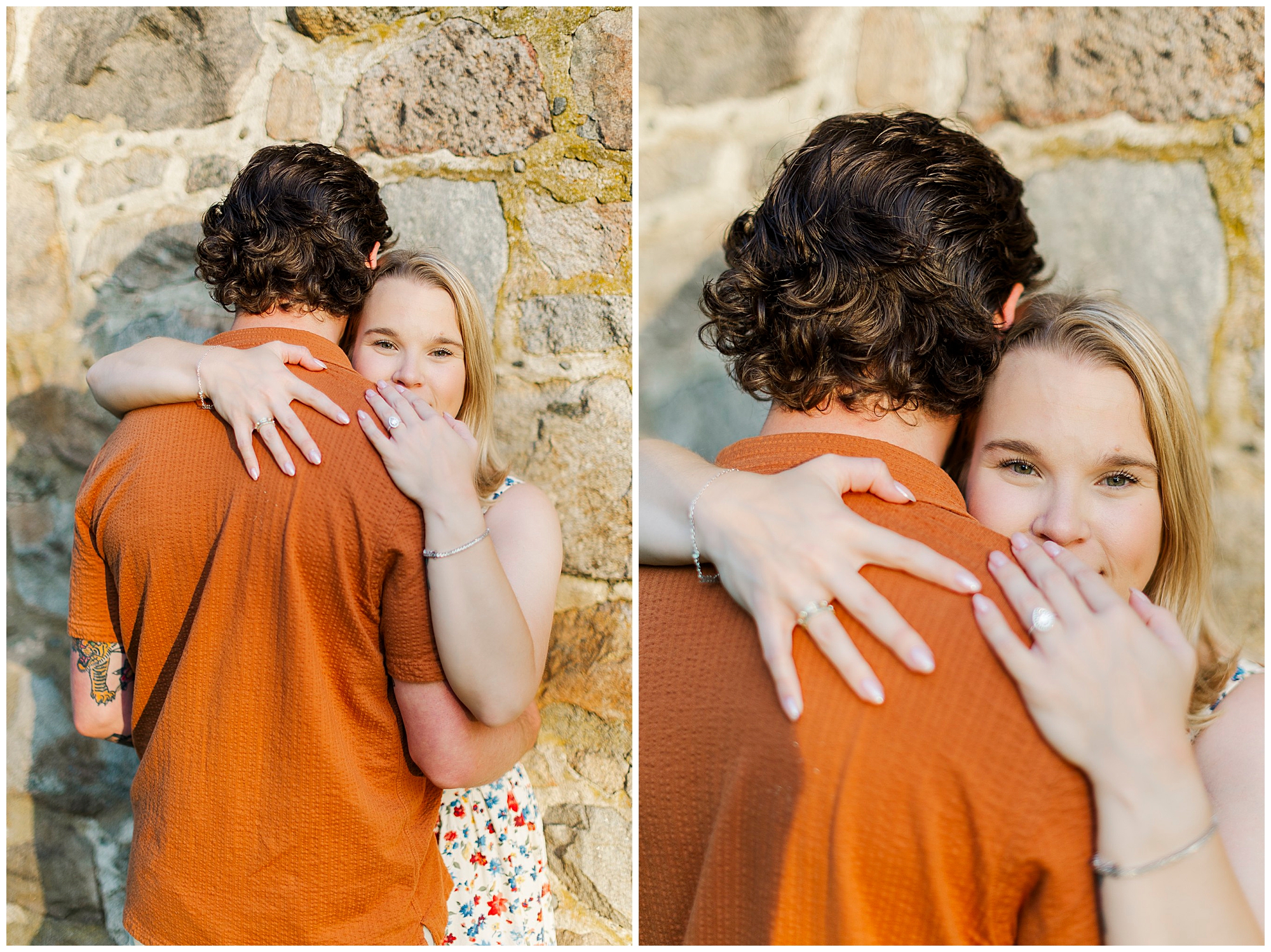 Woman in a floral dress embraces the man in an orange shirt and shows her engagement ring, smiling at the camera in front of a stone wall.