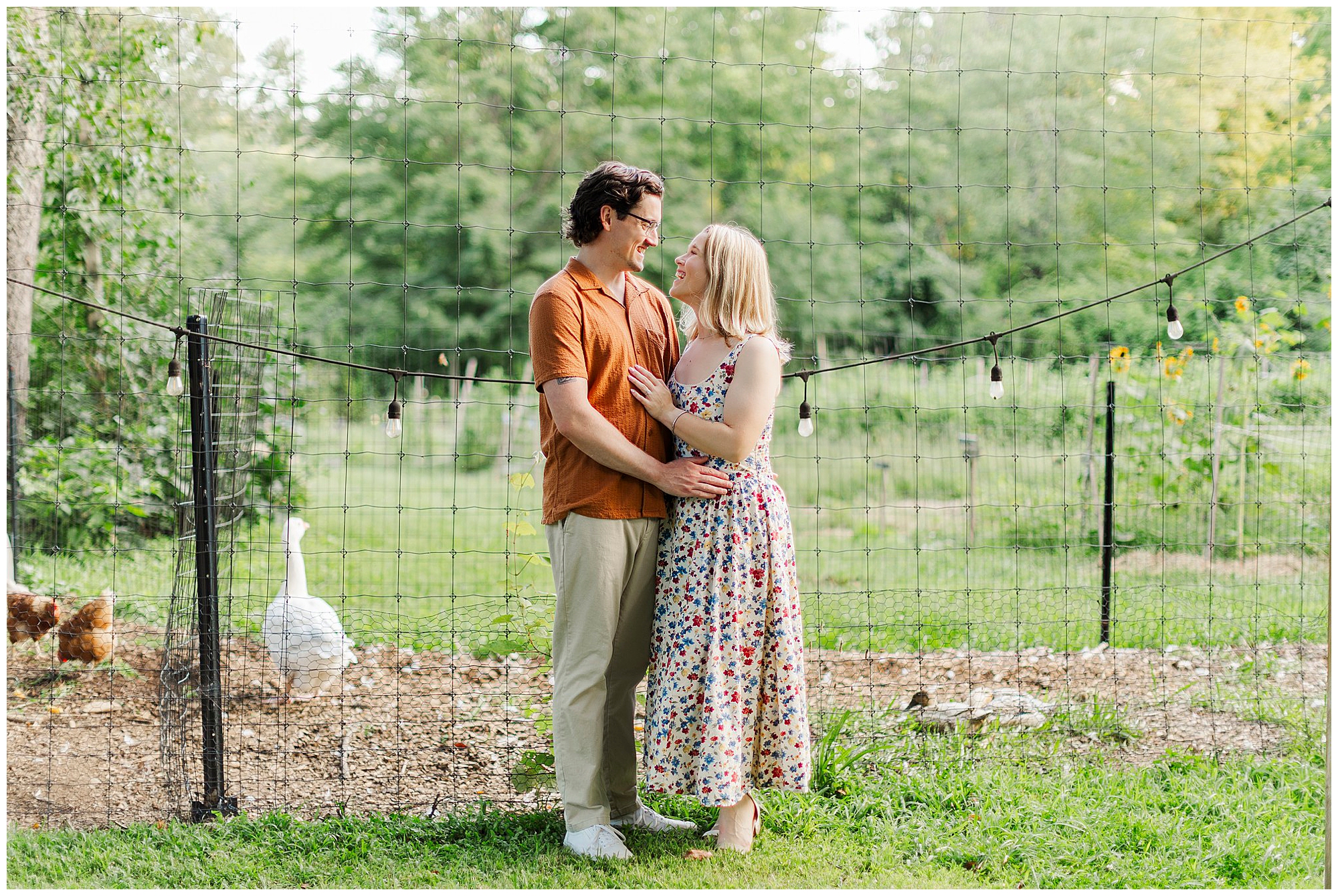 Couple embraces and smiles in a grassy yard with a wire fence, string lights, a goose and a few chickens behind them.