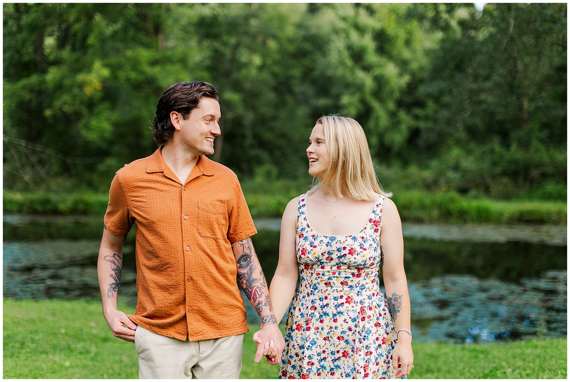 Couple holds hands and smiles at each other while walking near the pond, with lush green trees behind them.