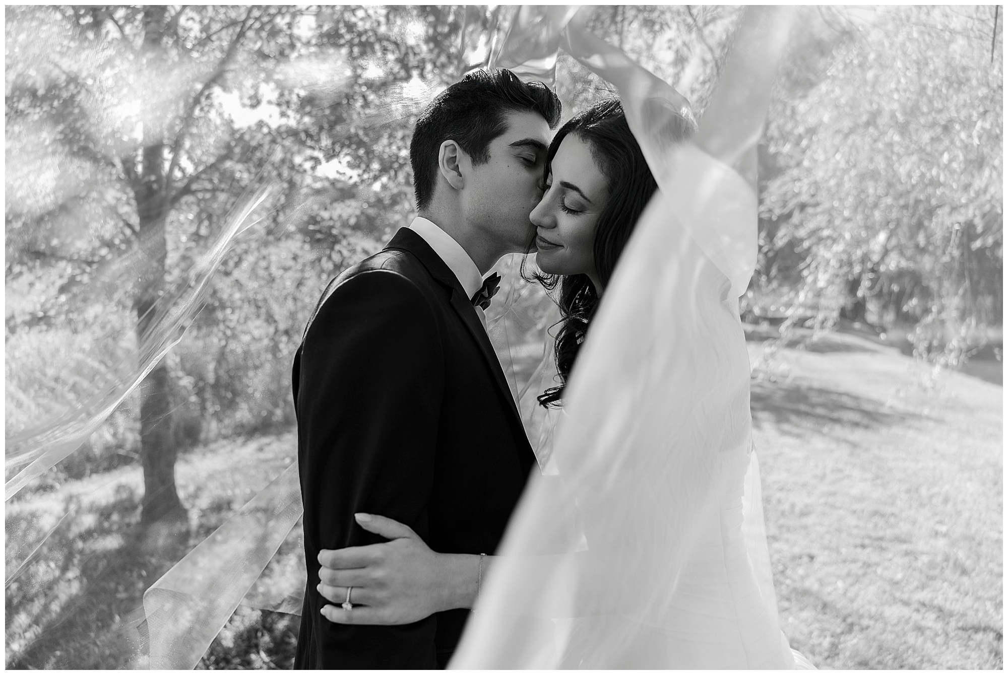 A groom in a suit kisses the bride’s forehead as she smiles with eyes closed, both embracing outdoors under sheer fabric, with sunlight filtering through trees at their intimate backyard micro-wedding.