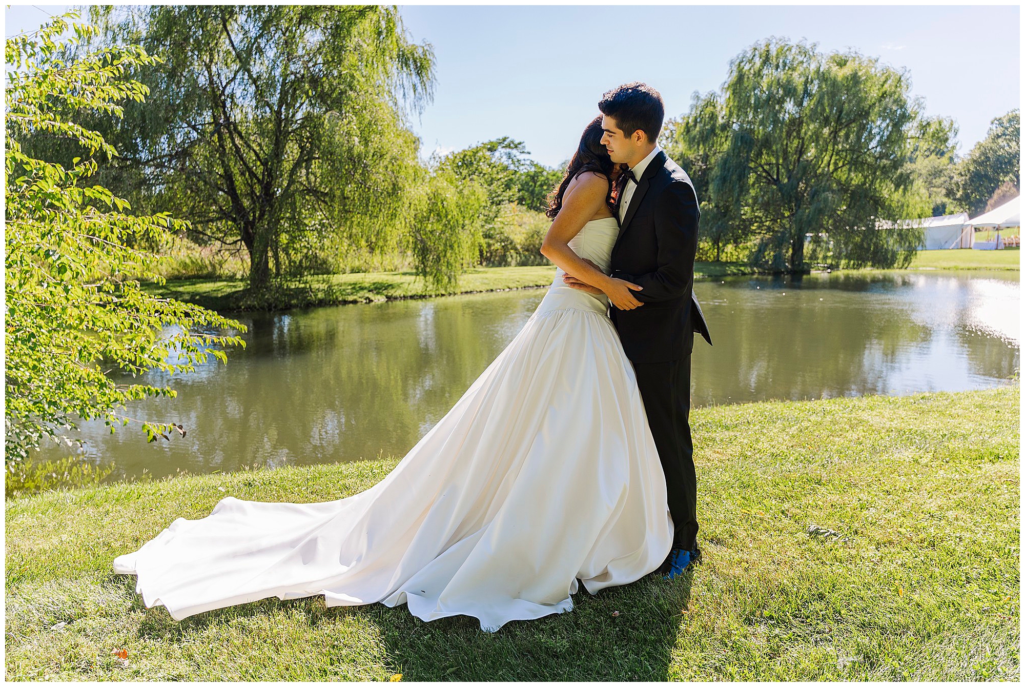 A bride and groom embrace by a pond on a sunny day, surrounded by green grass and trees during their intimate backyard micro-wedding. The bride wears a white gown, and the groom is in a black suit.