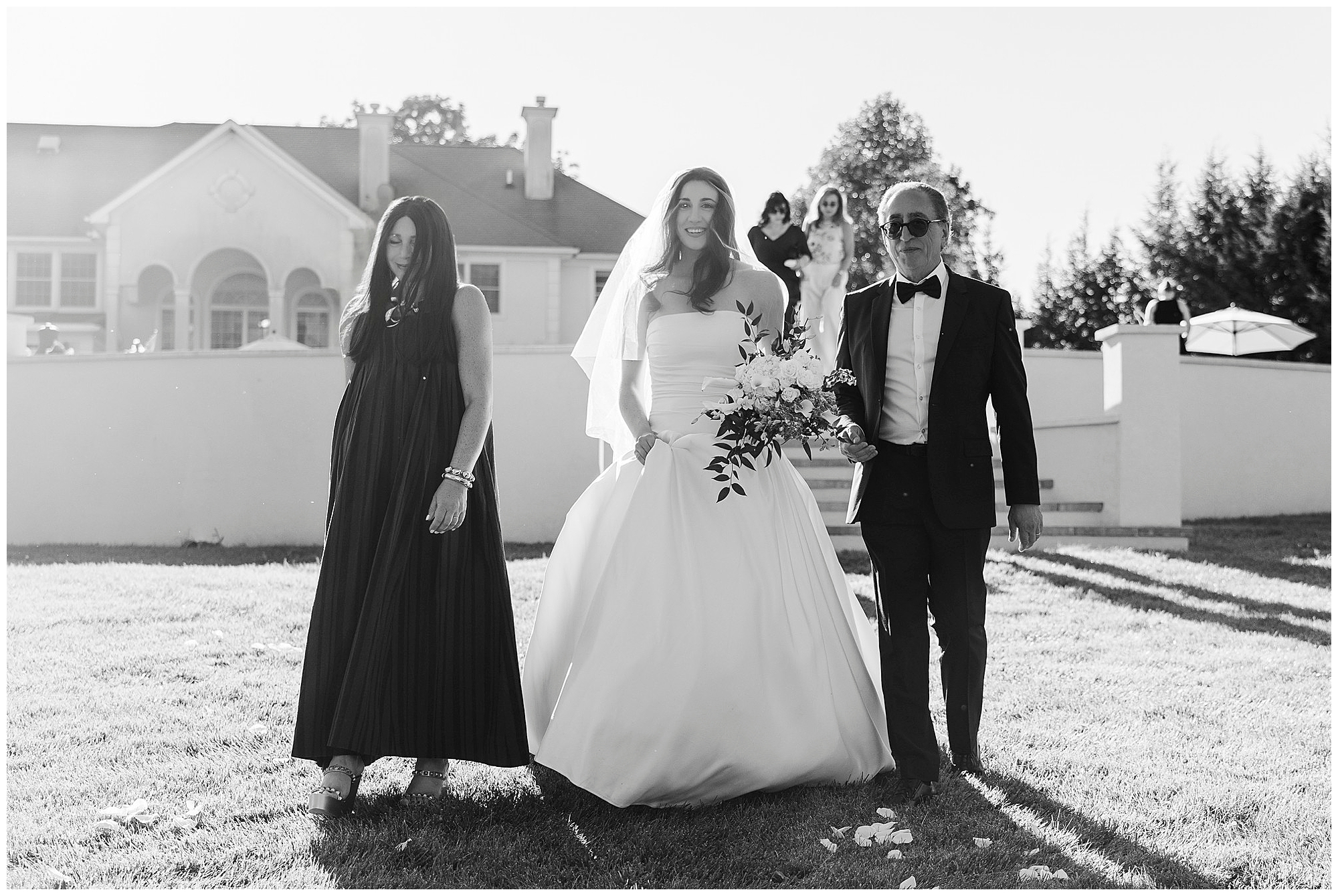 A bride in a wedding dress walks outdoors on grass, holding a bouquet, accompanied by a woman in a long dress and a man in a suit and sunglasses. Sunlight highlights them during their backyard micro-wedding, with a house and trees set beautifully in the background.
