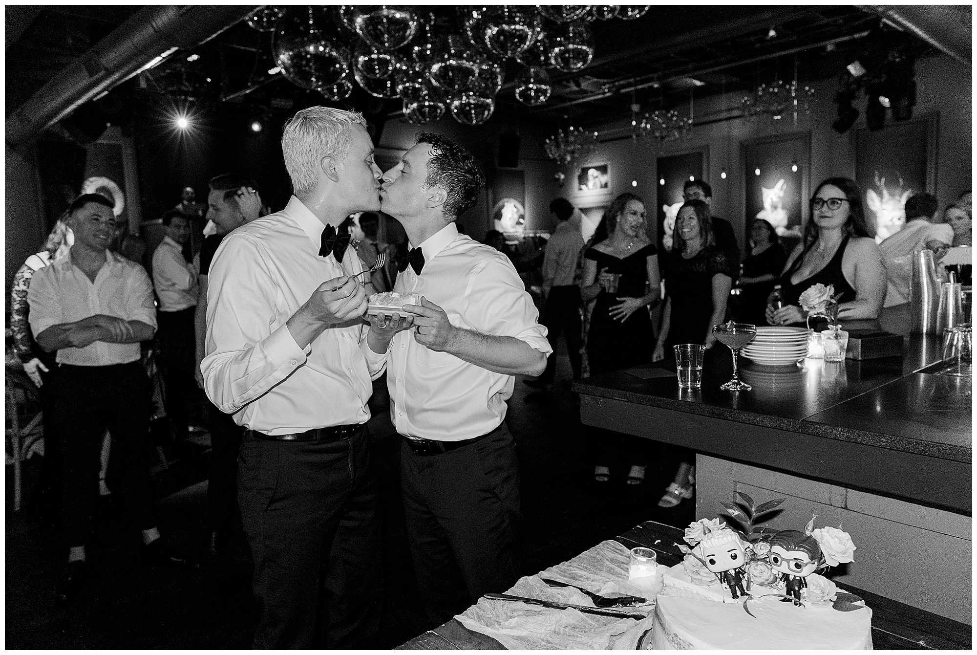 Two men in dress shirts and bow ties kiss while holding wedding cake at a lively Porta Asbury Park wedding reception, surrounded by smiling guests. The scene is joyful and celebratory, with a decorated cake in the foreground.