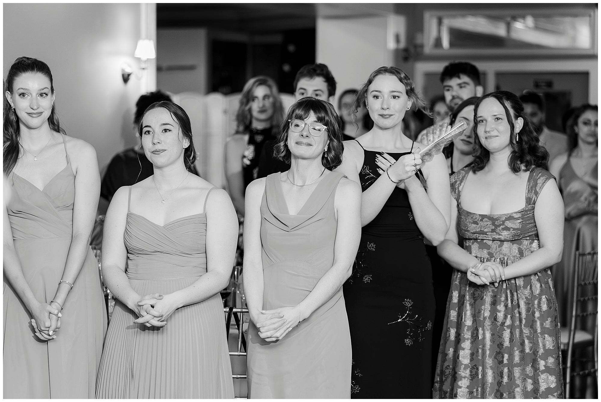 Group of women in formal dresses stand together smiling as they watch something out of frame.