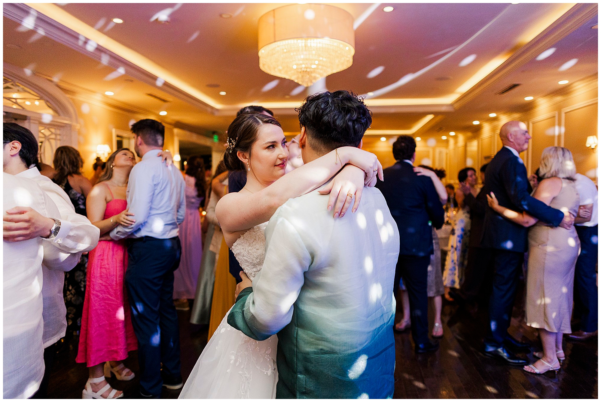 Bride and groom share a slow dance among their guests beneath warm lighting and a sparkling chandelier.
