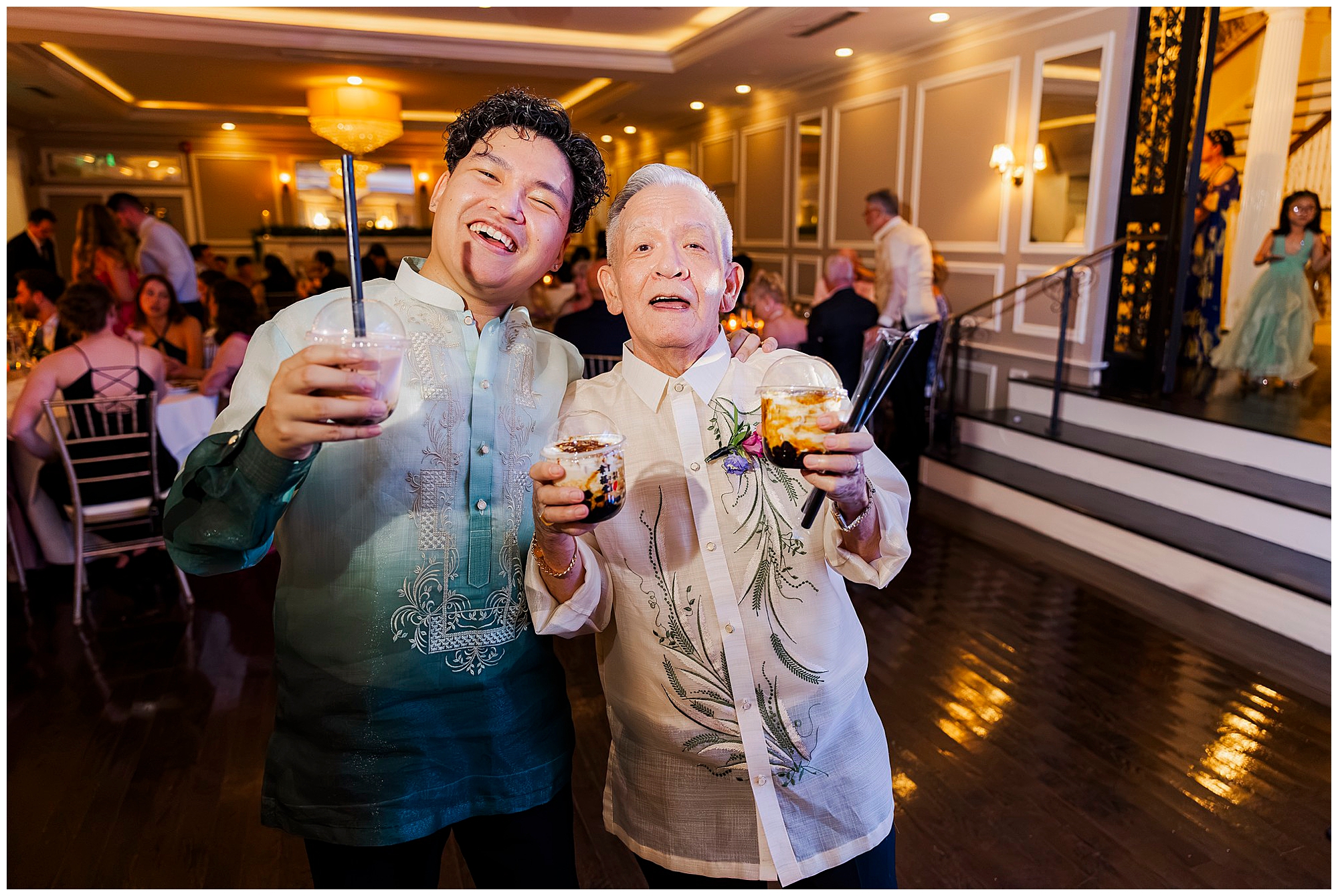 Two guests in traditional barongs smile and hold dessert drinks during the reception.