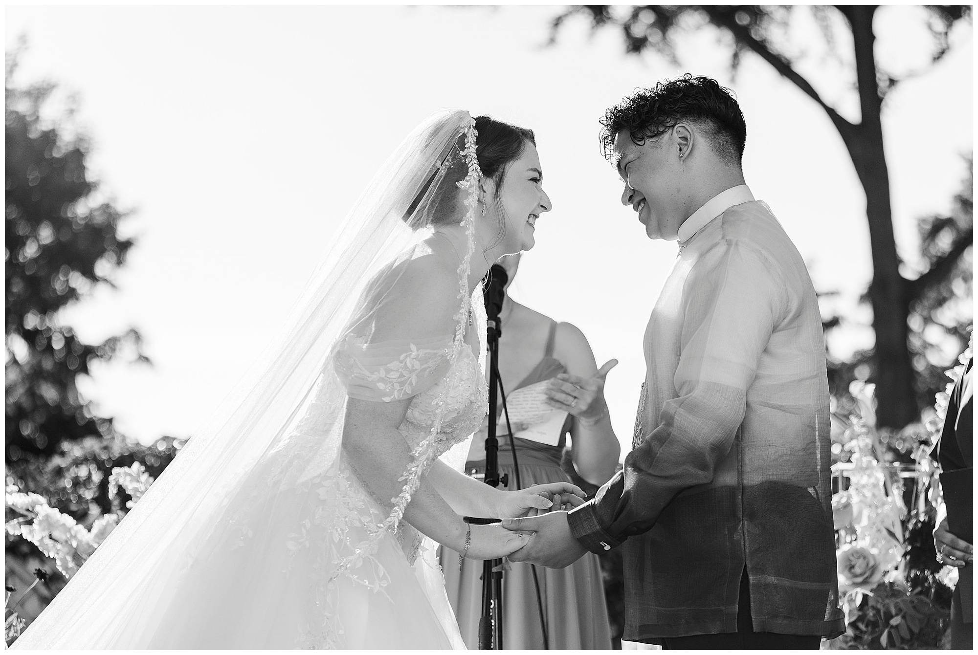 Bride in a veil and gown and groom in a Barong Tagalog smile and hold hands at the altar, a bridesmaid standing behind them.