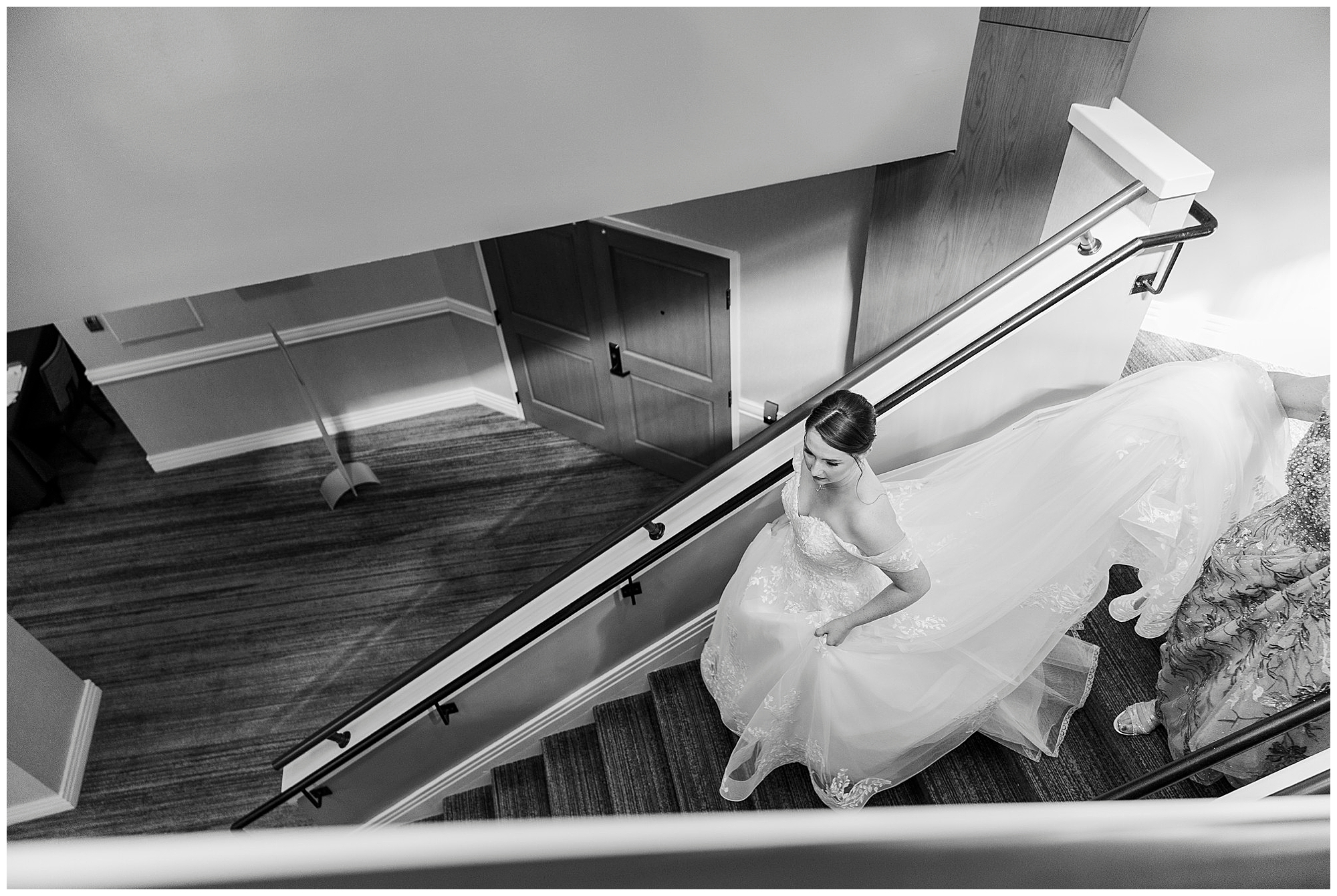 Black and white view from above of the bride climbing a staircase, holding her dress, with another person behind her.