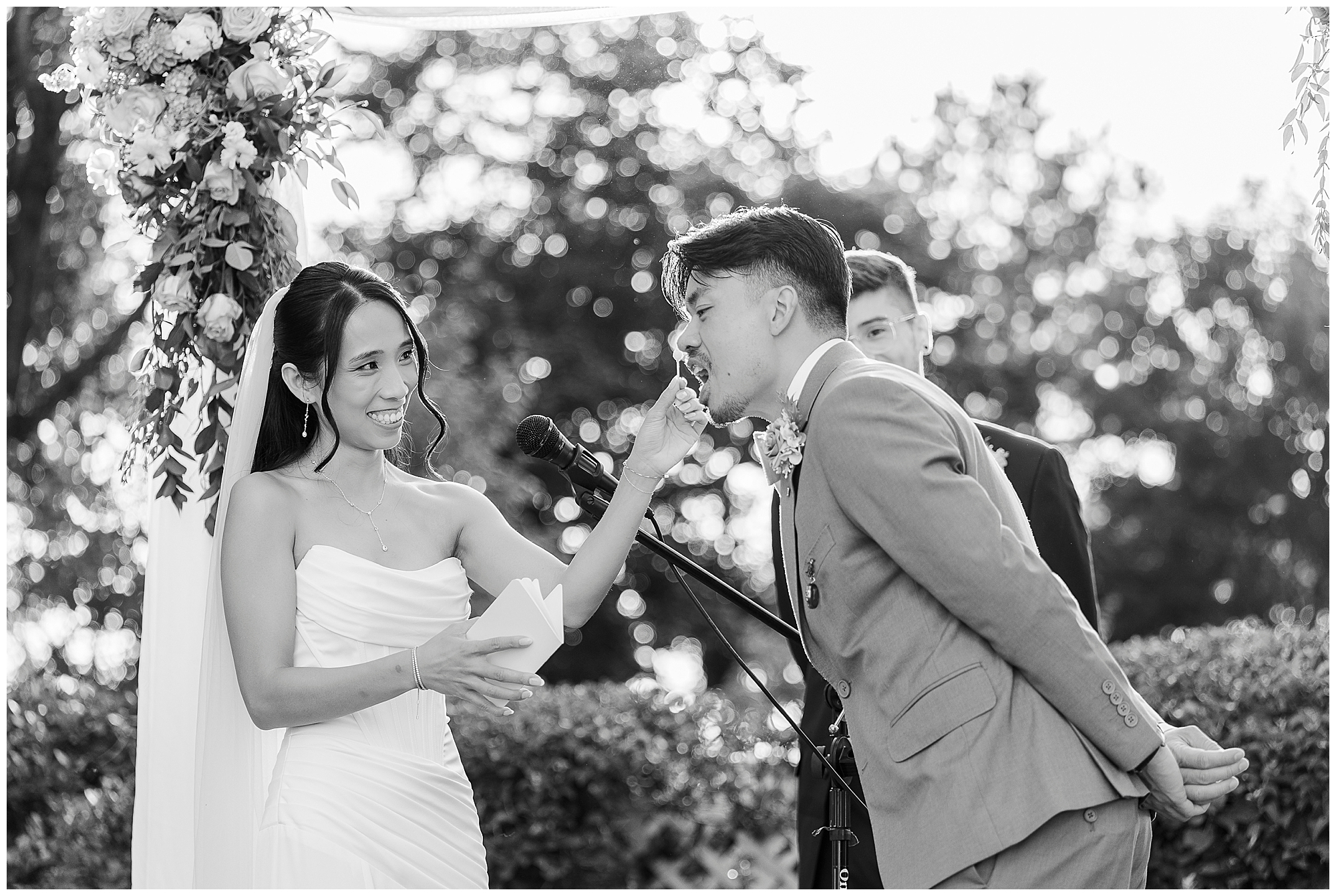 A bride in a white dress holds a note and feeds cake to a smiling groom in a suit during an outdoor wedding ceremony, with flowers and a microphone stand nearby.