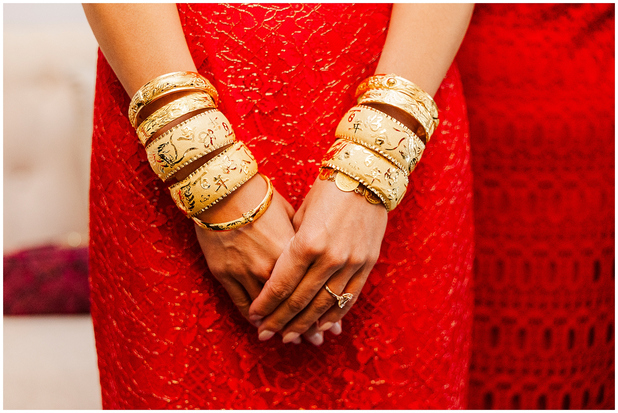 A person wearing an ornate red dress and multiple gold bangles stands with hands clasped. The focus is on the jewelry and dress, with a blurred background.