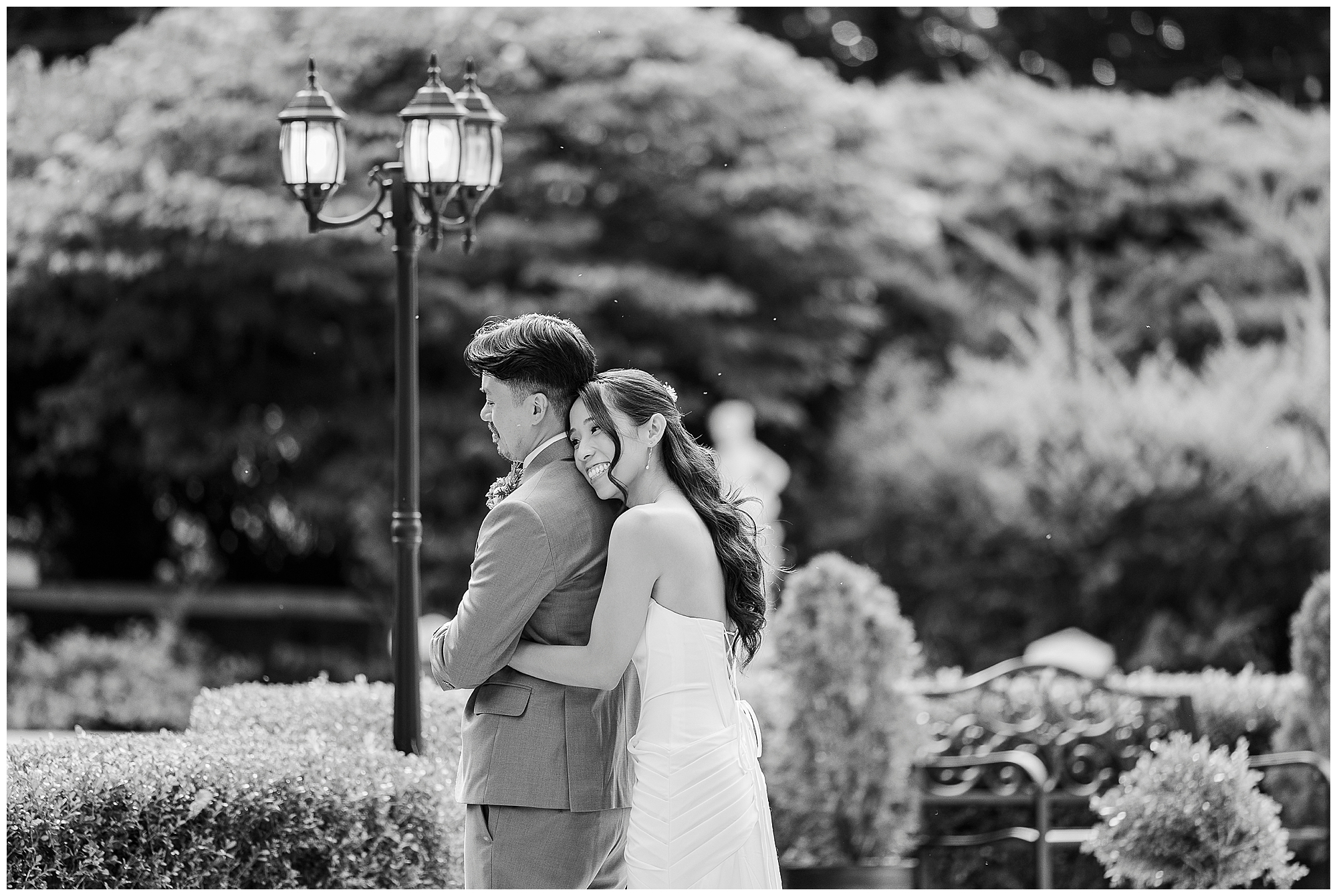 A woman in a white dress hugs a man in a suit from behind in a garden, both smiling. A vintage streetlamp and manicured bushes are visible in the background. The photo is in black and white.