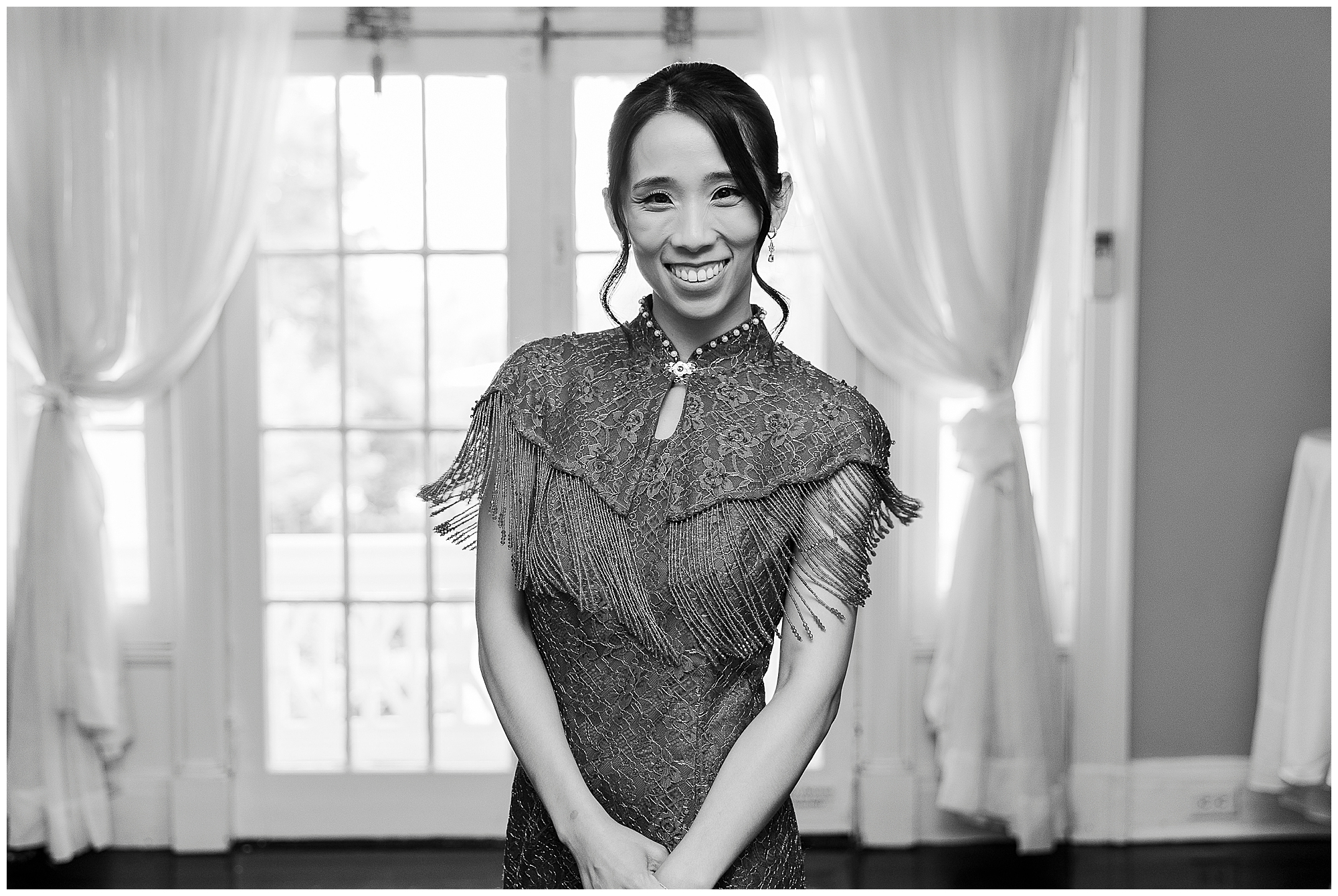 A woman in a lace dress with fringe stands smiling in front of tall windows with sheer curtains, her hands clasped in front of her.