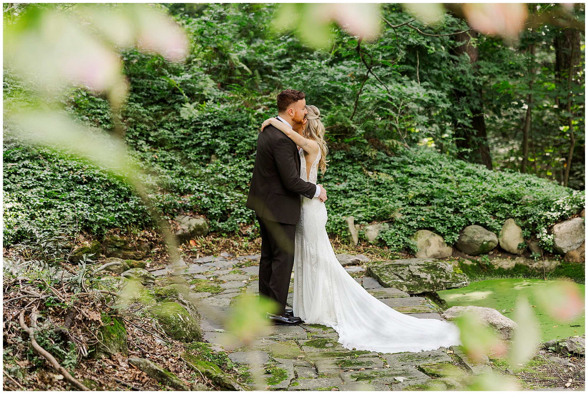 Bride and groom embrace on a stone path, framed by soft-focus leaves in the foreground.