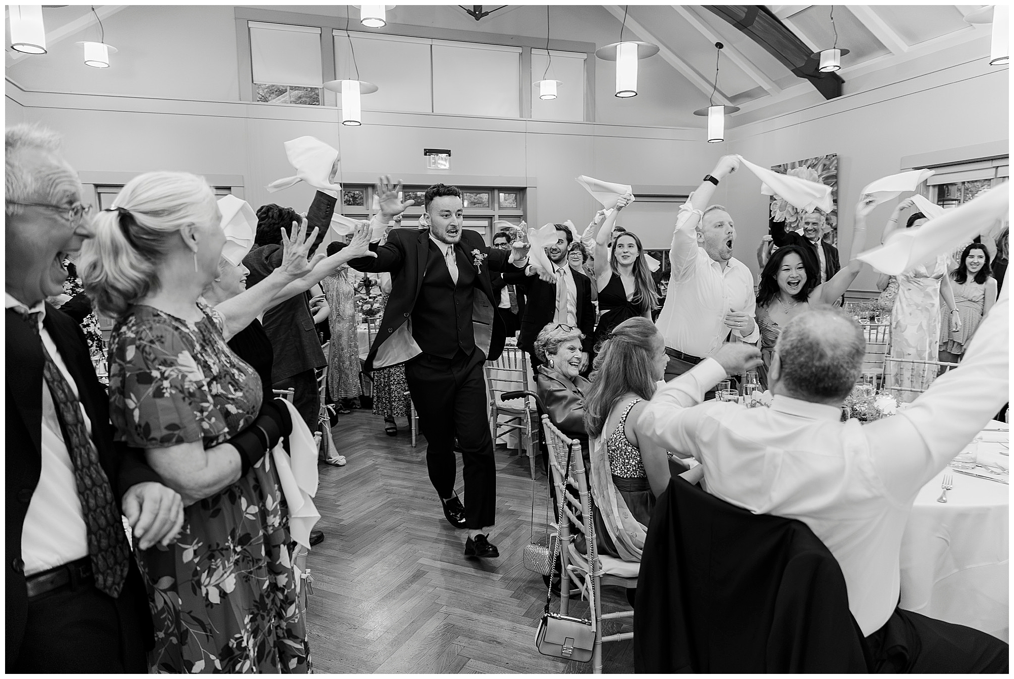 At a lively summer wedding reception, a group of people wave napkins in the air and cheer as a man in a suit walks energetically through the room. Everyone appears joyful and celebratory.