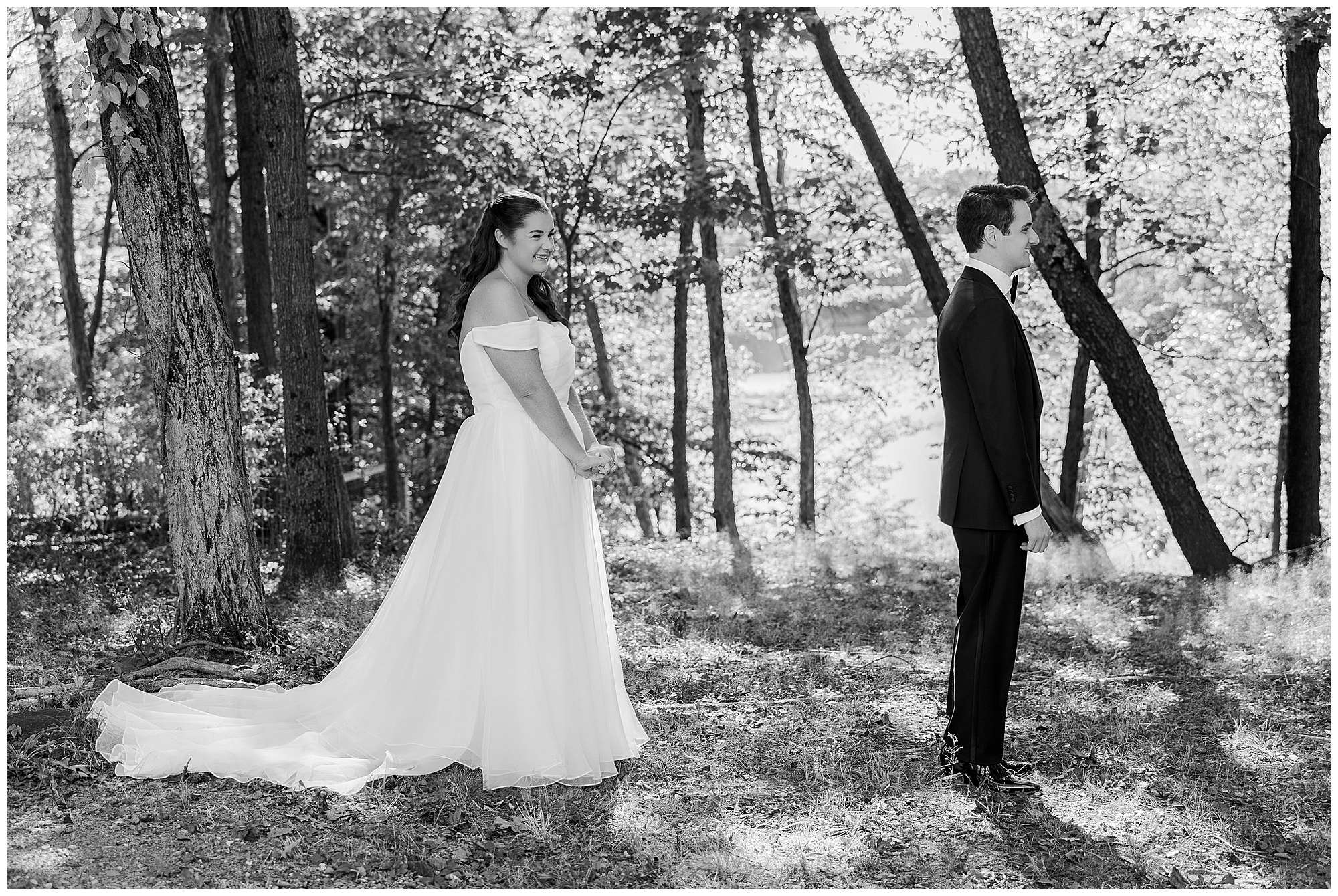 A bride in a long, flowing dress stands smiling behind a groom in a suit, both outdoors among trees, in a black and white photo.