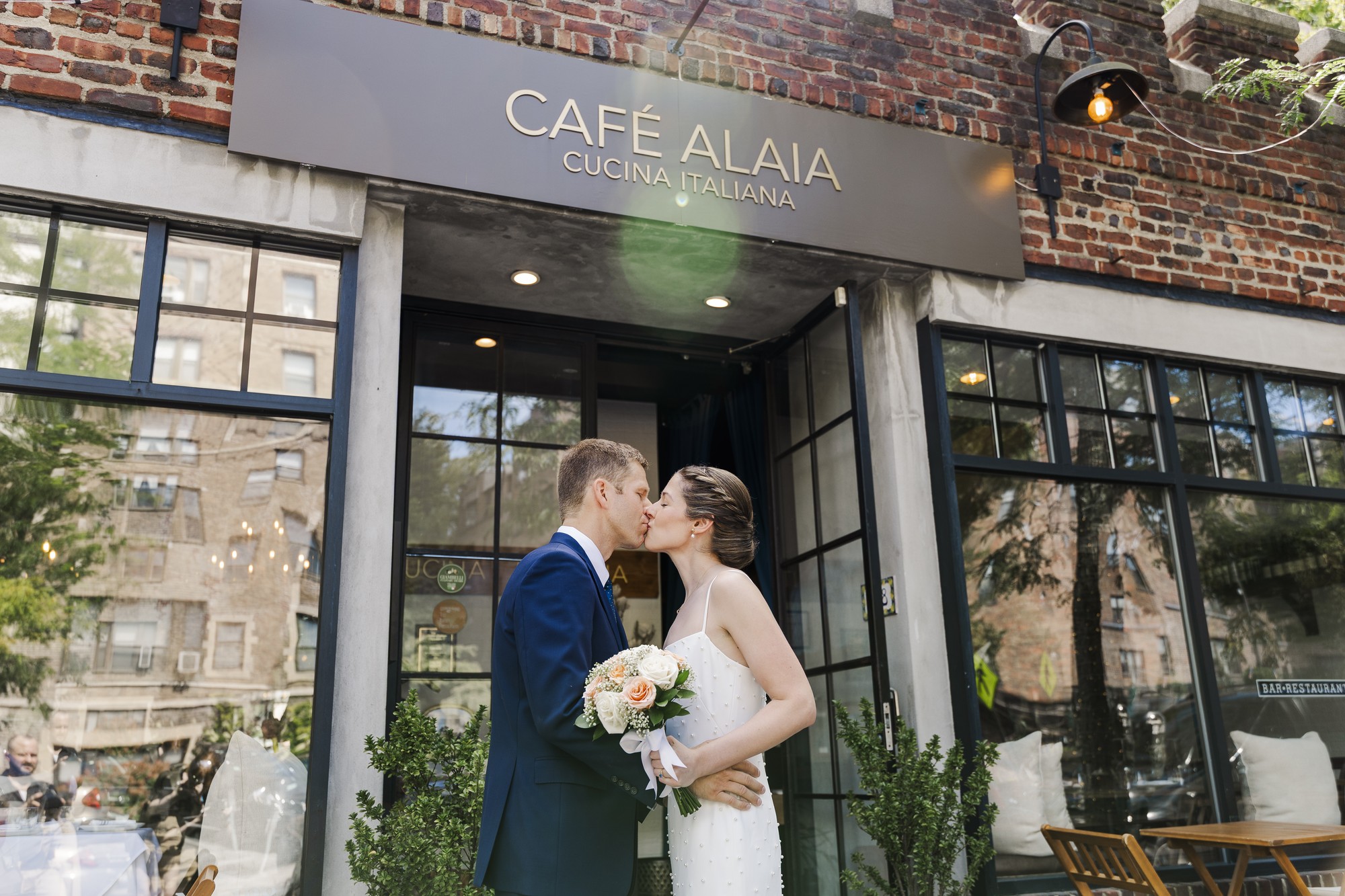 A bride and groom share a kiss outside Café Alaia following their elegant micro-wedding, with the bride holding a bouquet and both dressed in wedding attire.