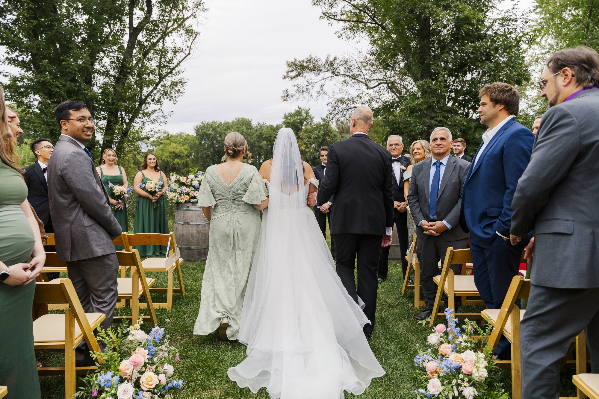 A bride walks down an outdoor aisle between rows of seated guests, accompanied by two adults. Wedding attendees smile, and floral arrangements decorate the aisle. Trees and greenery are visible in the background.