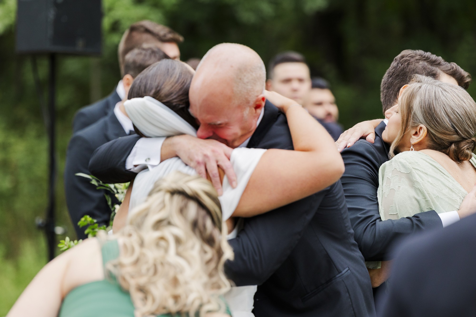 A group of people, dressed formally, embrace tightly outdoors, showing emotion and happiness. One man, appearing moved, hugs a woman in white while others join in the group hug. Green trees are visible in the blurred background.
