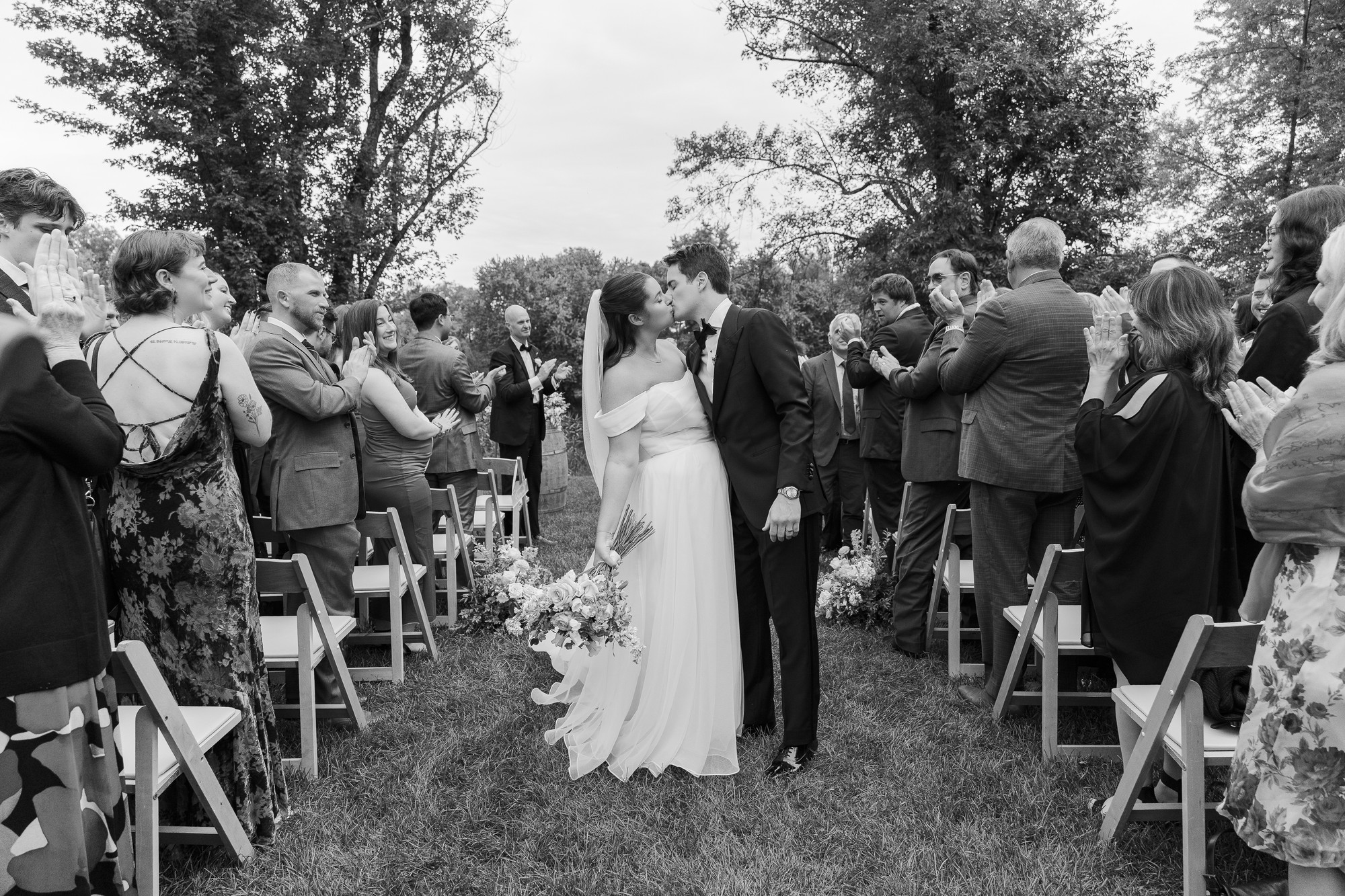 A couple in wedding attire kisses while walking down an outdoor aisle, surrounded by guests standing and applauding. The photo is in black and white, with trees and grass in the background.